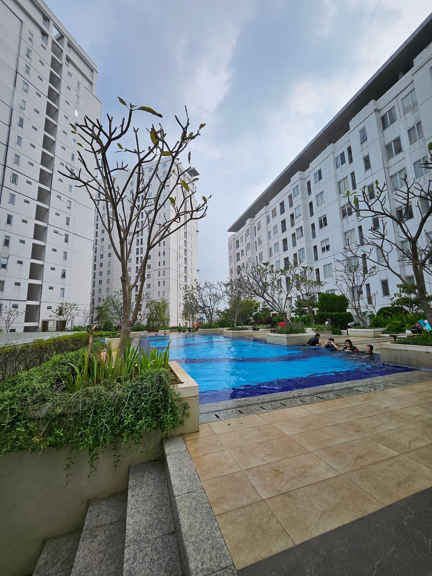 Modern apartment complex with a swimming pool and landscaped garden under a cloudy sky.