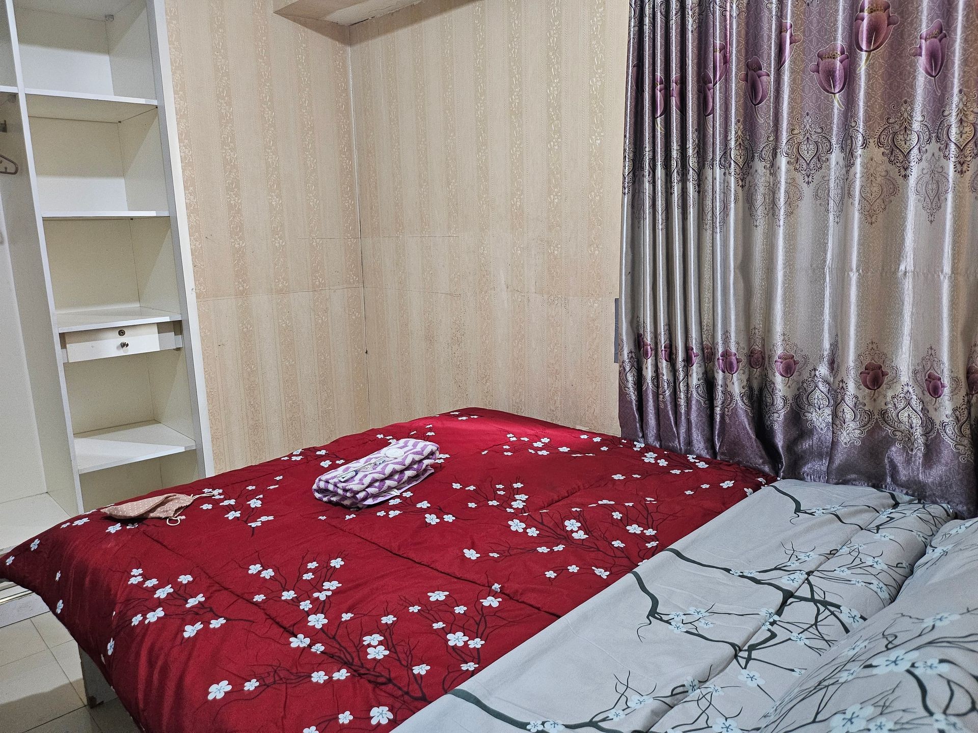 Bedroom with a red floral bedspread, closed curtains, and white shelves on the side.